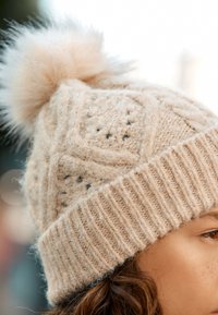 Knit beige beanie with a textured design, featuring a fluffy faux fur pom-pom on top and a ribbed cuff at the base.