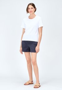 Biała bluzka z krótkim rękawem i okrągłym dekoltem, zestawiona z granatowymi szortami. Modelka nosi jasnobrązowe, plecione sandały. Minimalistyczny design, gładka faktura.