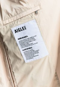 Μπεζ σακάκι με μια λευκή ορθογώνια υφασμάτινη ετικέτα ραμμένη στο εσωτερικό, που φέρει μαύρο κείμενο στα γαλλικά και τα αγγλικά κάτω από την επωνυμία AIGLE 1853.