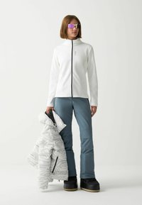 Giacca bianca con zip abbinata a pantaloni da sci blu. Una giacca puffer a righe grigio chiaro è tenuta in mano, completata da stivali invernali neri e occhiali da sole riflettenti.