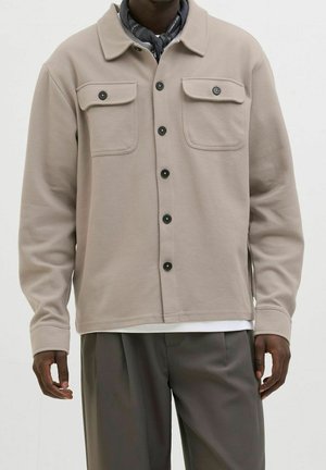 Mann trägt beige Hemdjacke mit Klappentaschen auf der Brust, dunkle Hose und ein dunkles Halstuch um den Hals, steht vor schlichter Hintergrund.