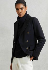 Blazer marine avec boutons dorés, design structuré et revers, porté sur un col roulé noir, complété par un pantalon crème.