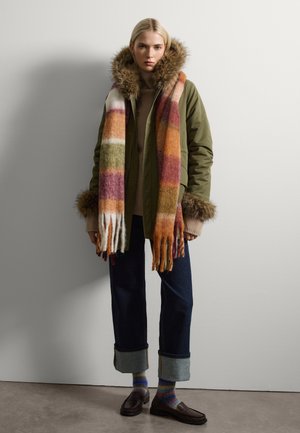 Parka verde oliva con capucha de pelo, suéter beige, jeans oscuros, bufanda de rayas multicolores y mocasines marrones con puños doblados.