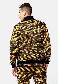 Mustard ja musta spordijakk, millel on bioohtlikkuse muster ja tekst "KARANTIIN". Materjal tundub sile ning seal on soonikkoes käe- ja kaelapaelad.