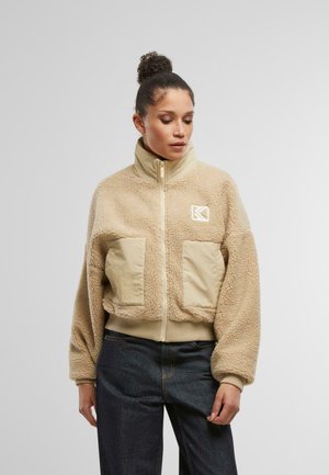 Beige Kunstpelzjacke mit hohem Kragen, Reißverschluss vorne, zwei Fronttaschen und elastischem Saum. Mit einem Logo auf der linken Brustseite.