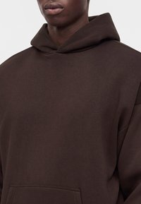 Gros plan d'une personne portant un sweat à capuche marron foncé uni avec une poche avant, montrant le cou, les épaules et une partie de la poitrine.