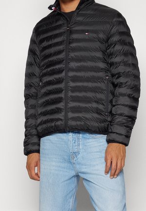 Mann trägt eine schwarze gesteppte Jacke mit Reißverschluss und Seitentaschen sowie hellblaue Jeans, Bild ist am Hals und an der Taille beschnitten.