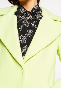 Long lime green coat with a collar over a black floral blouse featuring a tied neck detail; smooth texture and soft fabric.