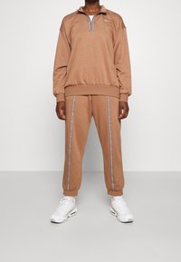 Brunt fleece atletisk set bestående av en halvmaskerad tröja med ribbade muddar och matchande byxor med sidostrapar. Bärs med vita sneakers.