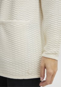 Sweter w kolorze kremowym, dziergany, z teksturowanym wzorem, długim rękawem, kieszonką na łaty oraz małym, okrągłym guzikiem na dole.