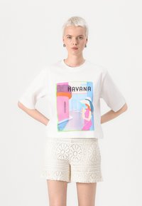 Balta medvilninė marškinė su spalvinga Havanos grafika ir geometriniais raštais. Modelis derina su kreminės spalvos nėriniuotomis šortais, parodančiais sudėtingus raštus.