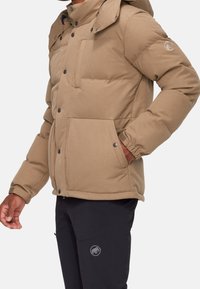 Béžová prošívaná bunda se zipem a zapínáním na patentky, nastavitelnou kapucí a dvěma předními kapsami; nošena přes tmavé kalhoty s logem.