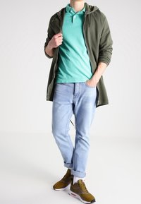 Felpa verde con zip sopra una maglietta polo teal, abbinata a jeans azzurri chiari e sneakers marroni con accenti bianchi.