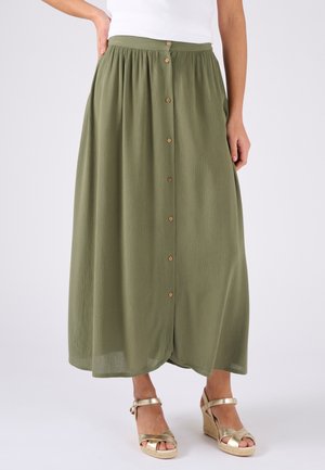 Falda midi verde oliva con tejido texturizado, que presenta un diseño en capas, botones en la parte delantera y una cinturilla. Combinada con sandalias de cuña doradas.