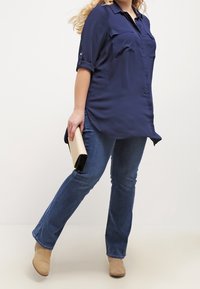 Camisa de botones azul marino con mangas remangadas, dos bolsillos en el pecho, combinada con unos jeans de mezclilla acampanados y botas de tobillo beige, sosteniendo un bolso de mano color crema.