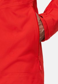 Giacca rossa con finitura testurizzata, tasca laterale con zip e polsini regolabili. Il materiale sembra leggero e resistente all'acqua.