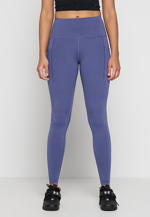 Femme portant des leggings violets taille haute avec poches latérales et chaussures de sport noires, debout devant un fond clair uni.