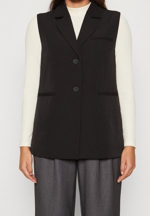 Femme portant un gilet noir boutonné sur un haut blanc à manches longues côtelé et un pantalon gris ample, debout les bras détendus.