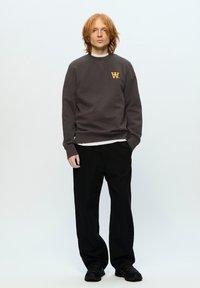 Dark gray sweatshirt with a round neckline, orange logo on the chest, paired with black pleated pants and black footwear.