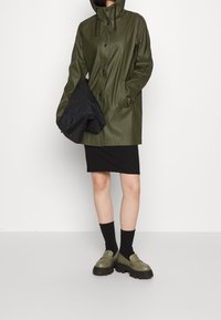 Giacca impermeabile verde oliva con cappuccio, bottoni frontali e vestibilità comoda, abbinata a una gonna plissettata nera e scarpe slip-on verdi.