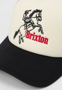 Cappello crema e nero con grafica ricamata di un cavaliere su un cavallo, con "Brixton" in testo rosso sulla parte anteriore.