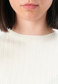 Gros plan sur le bas du visage et le cou d'une personne portant un pull blanc à côtes, avec des cheveux courts et foncés visibles au niveau du cou.