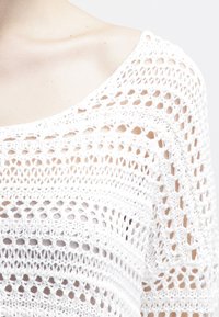 Haut tricoté blanc avec un motif en crochet ouvert, un large col et une coupe ample, agrémenté de textures variées et de rayures horizontales.