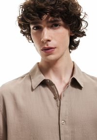 Beige button-up skjorte med et struktureret stof, kort krave og to knapper synlige foran, båret af en model med krøllet hår.