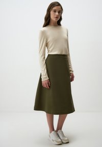 Olijfgroene A-lijn rok met een soepele textuur, gecombineerd met een crèmekleurig longsleeve shirt. Schoeisel: witte sportschoenen met beige accenten.
