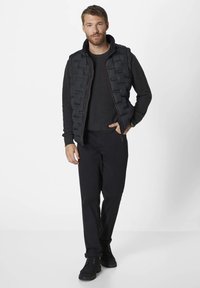 Schwarze gepolsterte Weste mit hohem Kragen und horizontalem Steppmuster, kombiniert mit einem dunklen Pullover und schwarzen Hosen, dazu Schnürstiefel.