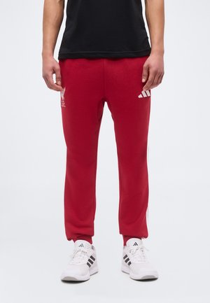 Personne portant un pantalon de survêtement rouge Liverpool FC avec des bandes blanches Adidas et des baskets blanches Adidas, debout devant un fond uni.