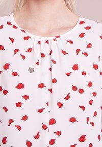 Blouse blanche avec un motif de petites casquettes rouges et une unique casquette grise, dotée d'un col rond et de tissu froncé au niveau du col.