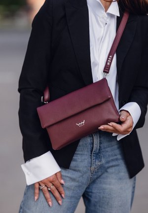 Bordó kožená kabelka přes rameno s přehnutým víkem, na kterém je stříbrné logo a nastavitelné ramínko, držena na černém saku.