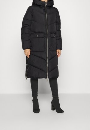 Osoba ubrana w czarną długą kurtkę puchową z przednim zamkiem błyskawicznym, kieszeniami z patkami oraz czarne botki na obcasie na jednolitym jasnym tle.