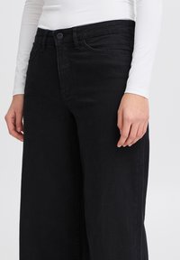 Jeans neri a gamba larga in denim, con vita media e chiusura a bottone, dotati di tasche frontali, esposti insieme a una maglietta bianca a maniche lunghe.