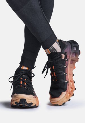 Ben gekruist, zwarte textuurleggings, oranje gestreepte sokken en zwarte trailhardloopschoenen met dikke, robuuste beige zolen.