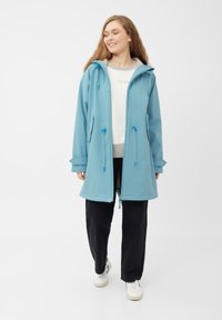 Giacca impermeabile azzurro chiaro con cappuccio, dotata di cordini regolabili, due tasche frontali e una vestibilità comoda. Abbinata a pantaloni neri e sneakers.