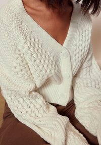 Stickad cardigan i off-white, med strukturerade ärmar, djup V-ringning och fyra stora knappar. Tyget verkar mjukt och chunky.