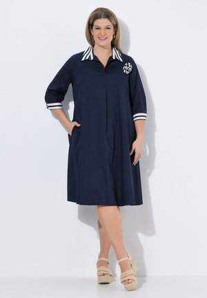 Smilende kvinde iført en marineblå knælængde kjole med stribet krave og ærmer, beige kilehælssandaler, stående mod en hvid baggrund.