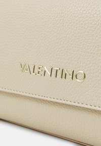 Beige nahkakotelo, jossa on teksturoitu pinta ja kullanvärinen "VALENTINO"-logo. Muotoilussa on näkyvissä saumat reunoilla.