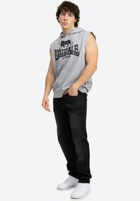 Gray sleeveless hoodie with a black logo, paired with black denim jeans and white sneakers. Features a front pocket and adjustable hood.