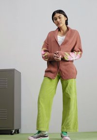 Cardigan de gran tamaño en color óxido con patrones florales rosas en las mangas, usado sobre una blusa blanca a rayas y combinado con pantalones de pana verde brillante.