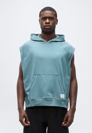 Junger Mann trägt eine ärmellose, türkise Kapuzenweste mit Fronttasche und schwarze Hose, steht vor einem schlichten, hellen Hintergrund.