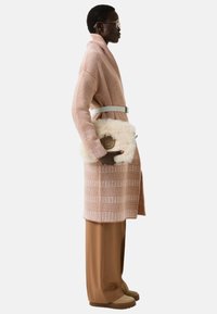 Woman in brown patterned coat with wide belt, matching wide-leg pants, beige boots, and a fluffy cream clutch bag, standing in profile.