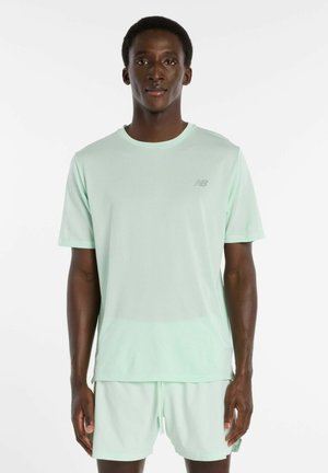 Mann, der ein hellmintgrünes New Balance Sportshirt mit kurzen Ärmeln und passende Shorts trägt und vor weißem Hintergrund steht.