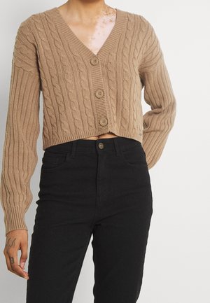 Person iført en kort, brun kabelstrikket cardigan med knapper og sort, høj taljede bukser, der viser en tatovering på venstre hånd.