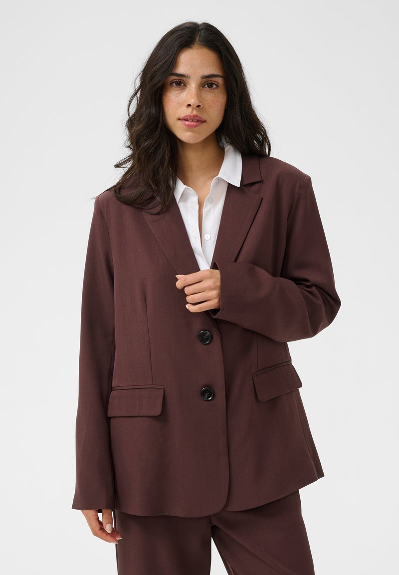 Femme aux longs cheveux noirs porte une veste de costume marron et un pantalon assorti par-dessus une chemise blanche boutonnée, debout devant un fond clair uni.