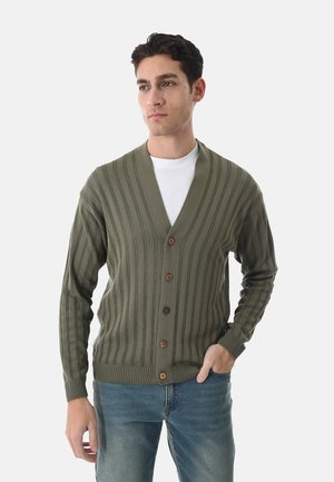 Uomo che indossa un cardigan verde oliva a coste con bottoni sopra una camicia bianca e jeans blu, in piedi con una mano in tasca, guardando di lato.