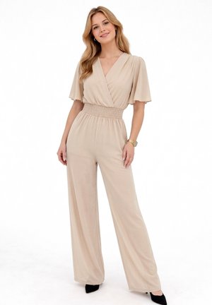 Kobieta o długich blond włosach, ubrana w beżowy kombinezon z szerokimi nogawkami i falbankowymi rękawami, w czarnych szpilkach z zaostrzonym noskiem, stojąca i uśmiechająca się.