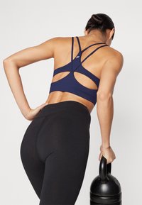 Navy sports bra with crisscross back design, paired with high-waisted black leggings, and holding a black kettlebell.